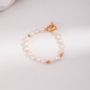 May include: A delicate bracelet featuring irregular, off-white pearls strung together. The bracelet has gold-colored accents and a gold-colored toggle clasp. The pearls are of varying sizes and shapes, creating a unique and elegant look.
