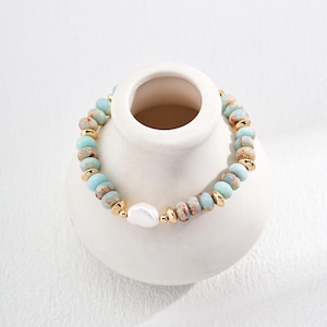 May include: A beaded bracelet featuring light blue and brown beads with gold-colored accents and a large white pearl. The bracelet is resting on a white ceramic vase, showcasing its design and colors.