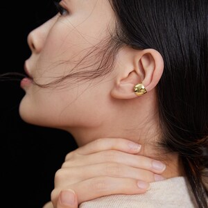 May include: A gold ear cuff with a textured, slightly curved design. The cuff is worn on the upper part of the ear.