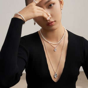 Op de afbeelding: Een vrouw draagt een zwart shirt met lange mouwen en een diepe V-hals. Ze draagt drie kettingen, een met een parelketting, een met een klein hanger en een met een grotere hanger.