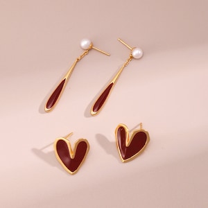 May include: A pair of gold earrings with red heart-shaped charms and a pair of gold earrings with a red teardrop-shaped charm and a white pearl.