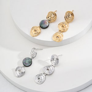 Op de afbeelding: Een paar oorbellen met een gouden en zilveren afwerking. Elke oorbel heeft drie ronde, getextureerde metalen stukken en een zwarte, ronde, iriserende steen.