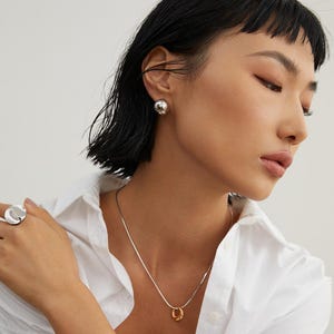 May include: A woman wearing a white shirt with a silver chain necklace and a gold ring pendant. She is also wearing silver earrings with a round design.