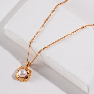 May include: A gold chain necklace with a textured square pendant featuring a white pearl.