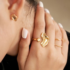 May include: A close-up of gold-colored jewelry, including a ring with an abstract, molten design, a delicate ring with small beads, and a matching earring. The jewelry is worn on a hand with white nail polish.