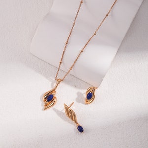 May include: A gold necklace with a blue gemstone pendant and matching earrings. The necklace has a delicate chain with small beads. The pendant and earrings feature a unique, abstract design.