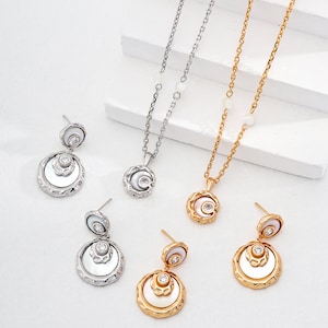 May include: Two sets of jewelry featuring a layered circle pendant and matching earrings. One set is silver with a white stone, the other is gold with a clear stone.