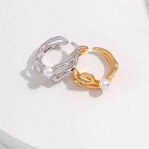 May include: Two open-ended rings, one silver and one gold, each featuring a small, round pearl. The silver ring has a layered design, while the gold ring has a more organic shape. Both rings are set against a white background.