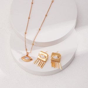 May include: A gold necklace with a small eye-shaped pendant and a pair of gold earrings with a square shape and dangling chains. The earrings have a clear stone in the center.