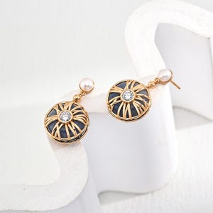 Op de afbeelding: Een paar gouden oorbellen met een blauwe steen en een parel hanger. De steen is omgeven door een gouden kooi ontwerp.