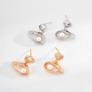 Op de afbeelding: Een paar oorbellen met een parel in het midden van een abstracte vorm van zilver en goud.