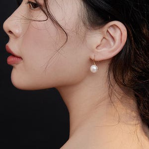 May include: A close-up of a woman's ear wearing a gold hoop earring with a white pearl dangling from it.
