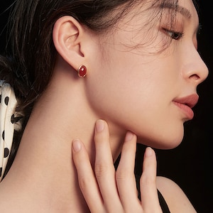 May include: A pair of gold stud earrings with a red oval gemstone. The earrings are worn by a person with long black hair.
