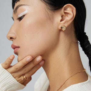 May include: A close-up of a person wearing a gold-toned earring with a textured, irregular shape. The earring is worn in the left ear. The person is wearing a white sweater and has dark hair.