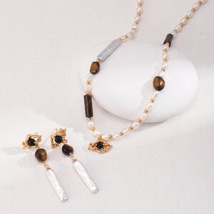 May include: A gold necklace with white pearls, tiger's eye stones, and a gold eye pendant. Matching earrings with black stones, tiger's eye stones, and white pearls.