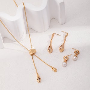 Op de afbeelding: Een gouden ketting met een geometrische hanger en twee hangende druppels. De ketting wordt gecombineerd met een paar gouden oorbellen, een met een geometrische druppel en de andere met een parel.
