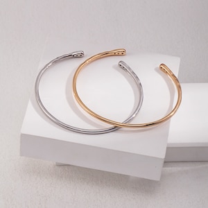 May include: Two simple bangle bracelets, one gold and one silver, with a hammered texture. The bracelets are open and have a slight curve to them.