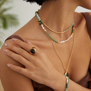 May include: A gold ring with a black oval gemstone. The ring is worn on the index finger of a hand. The hand is wearing a bracelet with green and white beads.