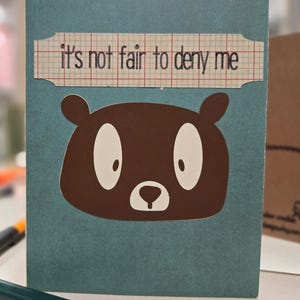 May include: A teal blue card with a brown bear face and the text "it's not fair to deny me" on a white and red gridded paper.