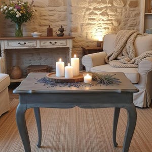 Pode incluir: Uma mesa de centro cinza, em estilo antigo, com pernas curvas, com velas e lavanda. A mesa está sobre um tapete tecido, com uma parede de pedra e um candelabro ao fundo. O quarto tem uma estética rústica e acolhedora.