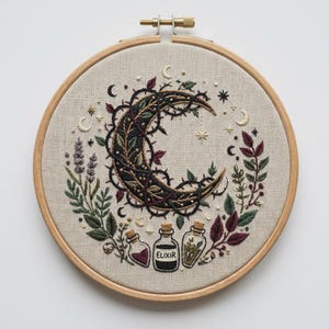 Peut inclure: Un tambour à broder en bois circulaire encadrant un tissu beige avec des motifs cousus à la main. Le centre présente un croissant de lune noir entrelacé de vignes épineuses, entouré de lavande, de feuilles, d'étoiles et de trois petites bouteilles en verre, dont une étiquetée ELIXIR.