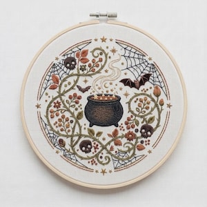 Peut inclure: Un tambour à broder circulaire contenant un panneau en tissu blanc avec un motif brodé à la main représentant un chaudron noir central dégageant de la fumée, entouré de vignes vertes, de feuilles brunes, de toiles d'araignée, de petites chauves-souris et de trois crânes.