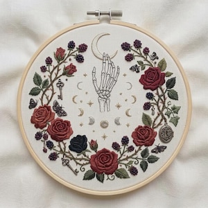 Peut inclure: Un tambour à broder contenant une pièce de tissu avec un motif cousu à la main représentant une main de squelette atteignant un croissant de lune. Le motif central est entouré d'une couronne de roses rouges, de mûres, de papillons, de clés et d'étoiles.