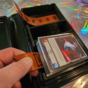 May include: Black, three-compartment card storage box with a stack of cards and orange dividers. The cards have text and graphics. The dividers have circular designs. The box is open, revealing the cards and dividers.