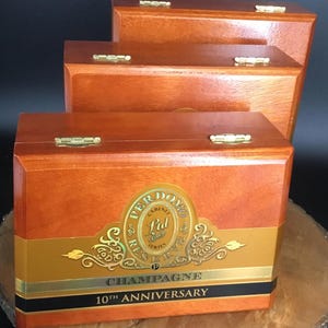 May include: Three rectangular wooden cigar boxes with a polished finish and gold-colored metal hinges. The front box features a prominent gold label with the text Perdomo Cabinet Series Ltd Reserve Champagne 10th Anniversary in black and gold.