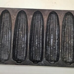 May include: A cast iron cornbread pan with six corn cob molds. The pan is dark gray and has handles on each end. The molds are detailed to resemble corn cobs.