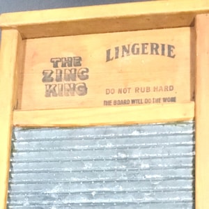 May include: A vintage wooden washboard with a corrugated metal scrubbing surface. The top section features the text "THE ZING KING" and "LINGERIE" in black lettering, with the phrase "DO NOT RUB HARD. THE BOARD WILL DO THE WORK."