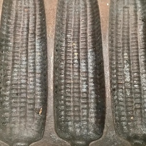 May include: Close-up of a cast iron cornbread pan with multiple corn cob molds. The pan is dark gray and shows signs of use. The detailed texture of the corn cobs is visible, suggesting a rustic, vintage style.