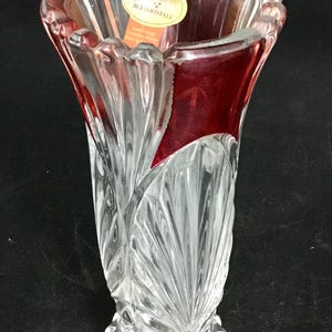 May include: A clear and ruby red lead crystal vase featuring a scalloped rim and a textured, fan-like vertical pattern along the body. An oval-shaped sticker with the text Nachtmann Bleikristall is attached to the interior rim of the glass vessel.