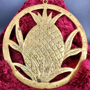 May include: A circular brass trivet featuring an openwork pineapple design with a textured, diamond-patterned fruit body and pointed leaves. The metal surface shows signs of age and wear, with a small hanging loop at the top center.