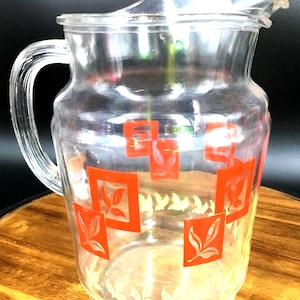 May include: A clear glass pitcher with a red and white geometric design. The pitcher has a wide mouth and a handle.