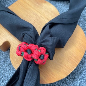May include: A black fabric scarf tied with a red and green paracord knot in the shape of two flowers. The knot is made with a paracord bracelet technique.