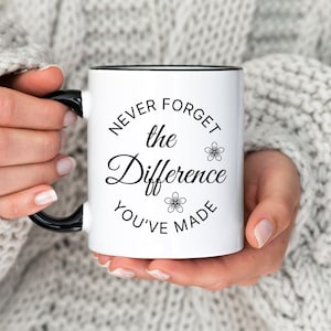 May include: White ceramic mug with a black rim and handle. The mug has a black graphic that says "Never forget the difference you've made" with small flower designs.