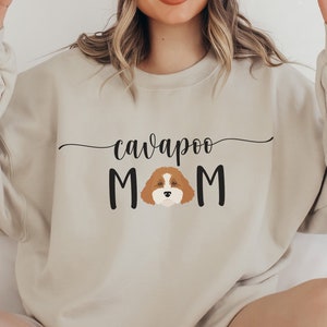 May include: A beige sweatshirt with the text "cavapoo MOM" in black lettering. A cartoon cavapoo dog is in the middle of the word "MOM".