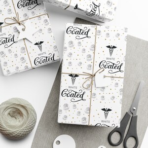 K&ouml;nnte beinhalten: Wei&szlig;es Geschenkpapier mit einem sich wiederholenden Muster aus goldenen Sternen und einer Schwarzwei&szlig;illustration eines Arztkittels und eines Caduceus. Der Text "WHITE Coated" wird auf dem Papier wiederholt.