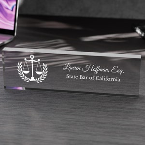 May include: A clear glass desk nameplate with a white scale of justice and laurel wreath design. The text "Lauren Hoffman, Esq. State Bar of California" is engraved on the nameplate.
