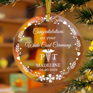 May include: A round, clear glass ornament with a white floral design and the text "Congratulations on your White Coat Ceremony" with the initials "PT" and a medical symbol. The ornament also includes the name "MADELINE" and the date "05-30-2024".