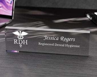May include: A clear acrylic desk nameplate with the name "Jessica Rogers" and title "Registered Dental Hygienist" in white lettering. The nameplate also features a white medical symbol and the letters "RDH".
