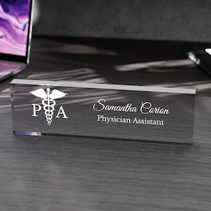 May include: A clear glass desk nameplate with a white medical symbol, the letters "PA", and the text "Samantha Corion Physician Assistant".