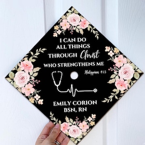 May include: Black graduation cap decorated with pink floral accents and the text "I CAN DO ALL THINGS THROUGH Christ WHO STRENGTHENS ME" and "EMILY CORION BSN, RN". A stethoscope and EKG line are also featured.