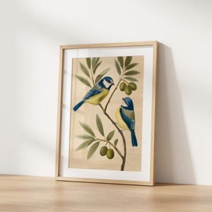May include: A framed wall art print featuring two blue and yellow birds perched on an olive branch with green leaves and small green olives. The illustration is set against a light beige background and enclosed in a light-coloured wooden frame.