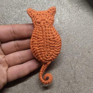 May include: A small, orange, crocheted cat-shaped applique featuring a textured yarn surface, pointed ears, a rounded body, and a long, curled tail. The piece is crafted with visible yarn loops and stitches, forming a solid, flat, cat-like silhouette.