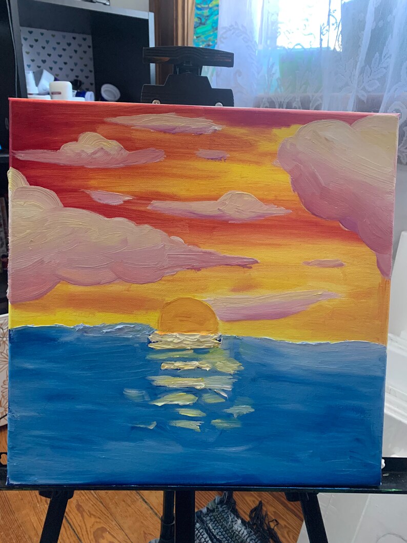 Pode incluir: Uma pintura de um p&ocirc;r do sol sobre o oceano. O c&eacute;u &eacute; uma mistura de vermelho, laranja e amarelo, com nuvens cor-de-rosa e fofas. O sol se p&otilde;e atr&aacute;s do horizonte, refletindo na &aacute;gua azul.