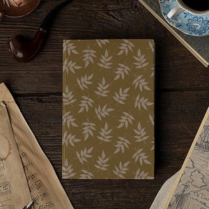 Può includere: Una natura morta con un diario con motivo a foglie, una tazza di caffè, una pipa, una candela e carte antiche. Il diario ha uno sfondo dorato con un motivo ripetuto di foglie grigio chiaro. La tazza di caffè è blu e bianca.