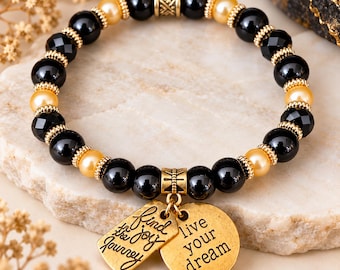 Op de afbeelding: Een kralenarmband met afwisselend zwarte en goudkleurige kralen en goudkleurige tussenstukjes. Twee goudkleurige metalen bedels hangen in het midden, een rechthoekige met de tekst 'find the joy in the journey' en een ronde met 'live your dream'.