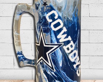 Op de afbeelding: Een bierpul van helder glas met een wervelend blauw en wit gemarmerd verfontwerp. De pul toont een grote marineblauwe ster met een witte omlijning en het woord COWBOYS verticaal gedrukt in dikke witte hoofdletters.